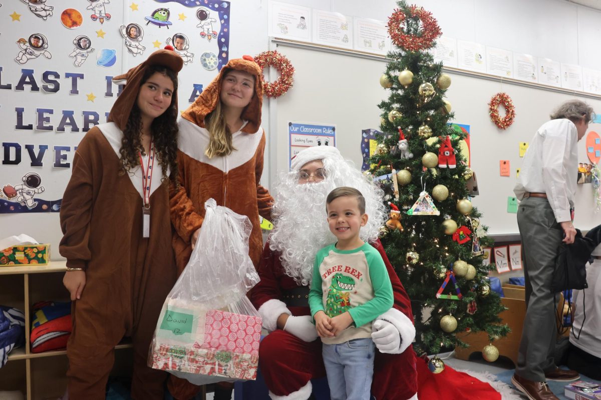Eighth graders Daniel Sanchez, Stella Roubach-Tomeo, and Sofia Queen pose to take a photo with their kid. As Santa, Sanchez delivers his gift to the kid.